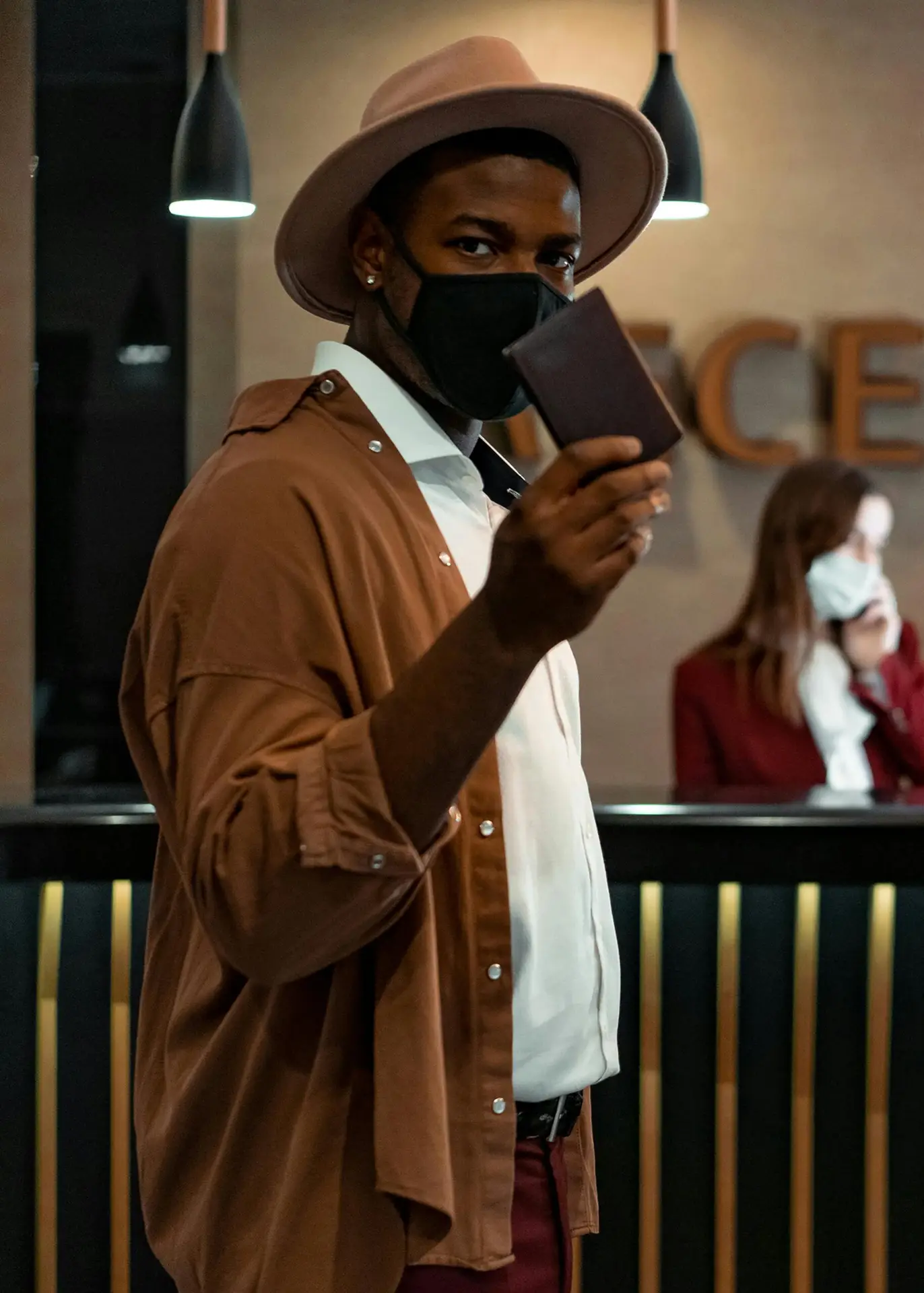 A man wearing a mask and hat holds a passport at a hotel reception, emphasizing travel safety.